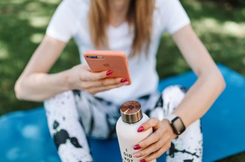 Woman using smartphone while holding bottle on yoga mat outdoors.
